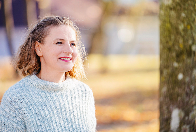 Alexandra steht im Wald lächelt in die Ferne. Im Hintergrund sind Bäume zu sehen.