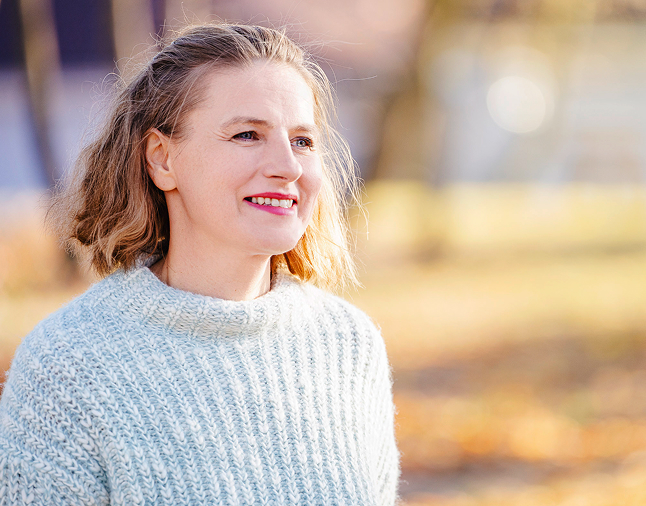 Alexandra steht im Wald lächelt in die Ferne. Im Hintergrund sind Bäume zu sehen.